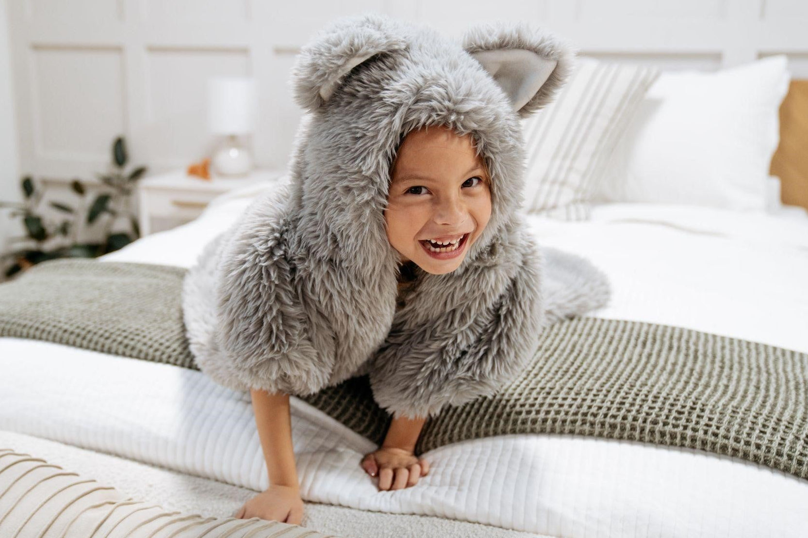 Child wearing a gray furry costume with wolf ears on a bed.